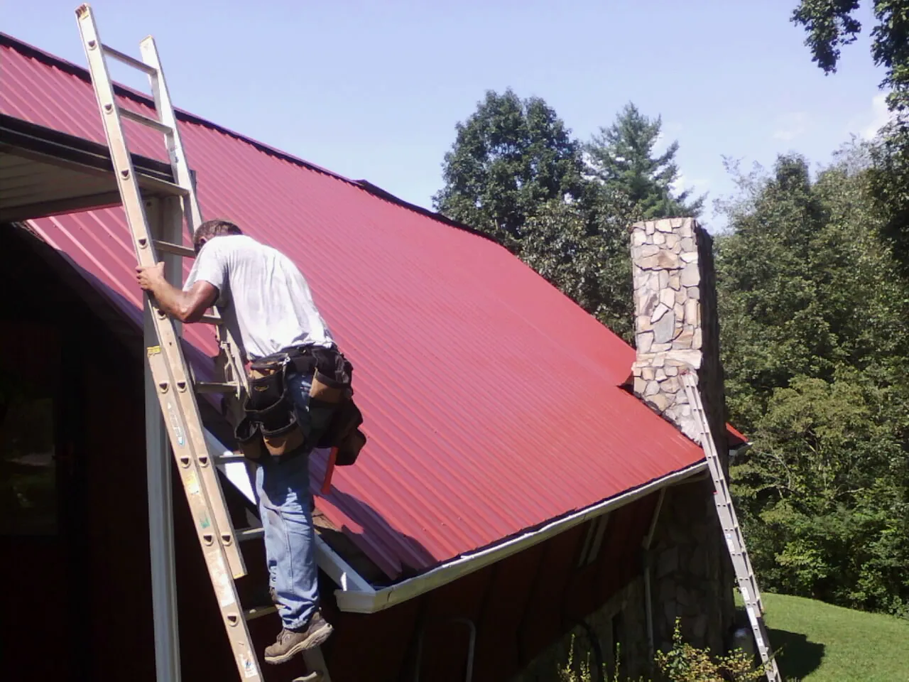 Roofer on a ladder beside a red metal roof and stone chimney — Soffit & Fascia Repair by Roofing Brothers in Theodore